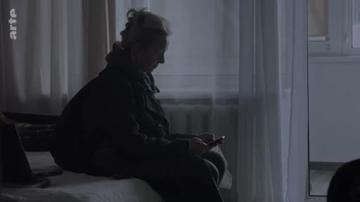 Une femme est assise sur un lit dans une chambre plongée dans la pénombre, utilisant son téléphone portable. Cette scène illustre l'isolement ou la communication numérique dans un contexte domestique.