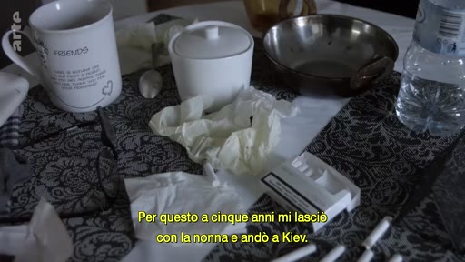Une vue en plongée d'une table de cuisine avec une tasse, un sucrier, un bol et un paquet de cigarettes, accompagnée de sous-titres en italien évoquant un départ pour Kiev.