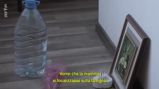 Un plan fixe montre un grand bidon d'eau en plastique et un cadre photo posé sur un sol en parquet, accompagné de sous-titres en italien évoquant la famille.