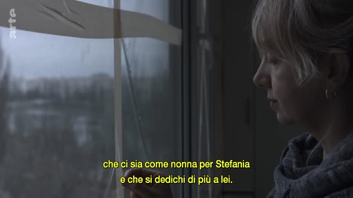 Une femme, vue de profil, regarde par la fenêtre dans un intérieur domestique. Le sous-titre à l'écran mentionne le rôle de grand-mère pour Stefania.
