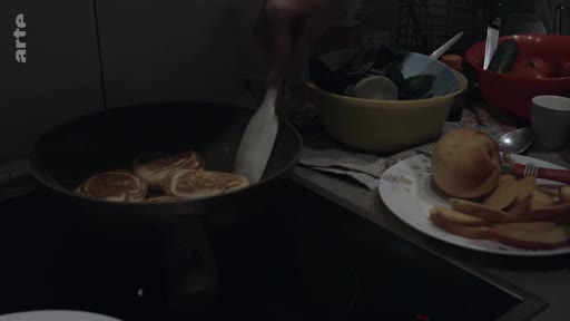 Une personne cuisine des pancakes dans une poêle sur une plaque de cuisson, avec des fruits tranchés sur une assiette à proximité, illustrant une routine domestique.