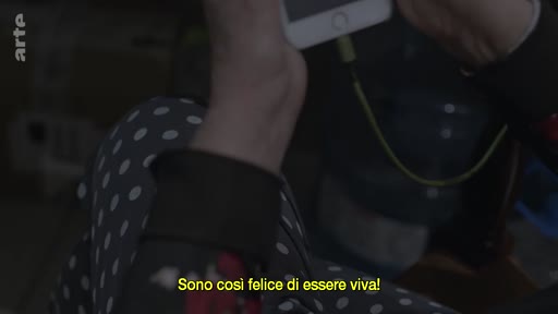 Une personne âgée, vêtue d'un pantalon à pois, tient un smartphone pour un appel vidéo. Le sous-titre italien indique 'Sono così felice di essere viva!' (Je suis si heureuse d'être en vie !).