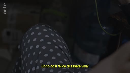 Une personne âgée, vêtue d'un pyjama à pois, participe à un appel vidéo et exprime son bonheur d'être en vie. L'image montre une interaction personnelle et émotionnelle capturée via un écran.