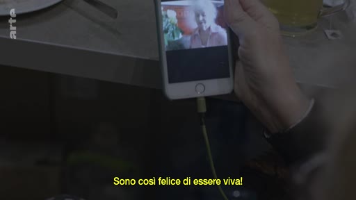 Une personne tient un smartphone montrant une femme âgée souriante lors d'un appel vidéo, avec le sous-titre italien 'Sono così felice di essere viva!' affiché à l'écran.