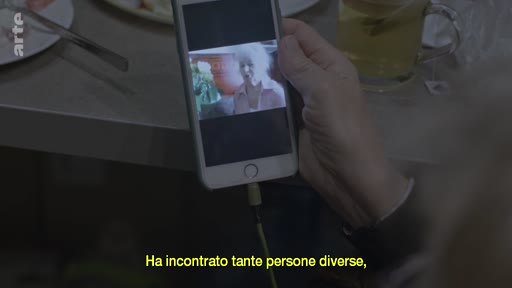 Une personne tient un smartphone montrant une femme âgée lors d'un appel vidéo, illustrant le thème de la communication à distance et des relations familiales. Le sous-titre italien indique qu'elle a rencontré beaucoup de personnes différentes.