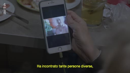 Une personne communique en appel vidéo avec une femme âgée, visible sur l'écran d'un smartphone. L'image est accompagnée de sous-titres en italien.