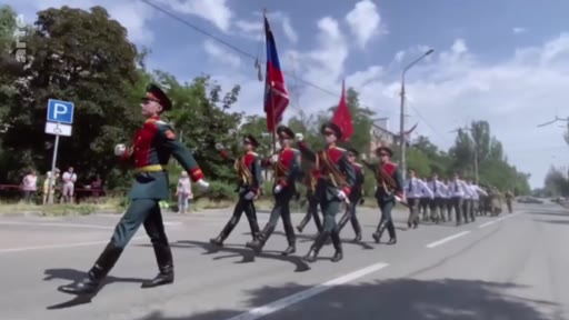 Des soldats en uniforme de parade défilent dans une rue de Marioupol, portant des drapeaux russes. Cette scène illustre la présence militaire russe dans la ville ukrainienne occupée.