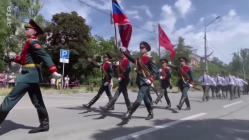 Des soldats en uniforme de parade défilent dans les rues de Marioupol, portant le drapeau russe et le drapeau soviétique. Cette scène illustre l'occupation de la ville ukrainienne par les forces russes lors d'une cérémonie militaire.