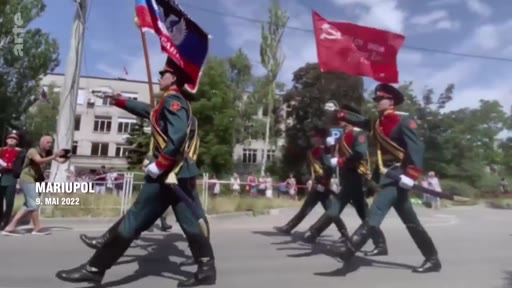 Des soldats russes défilent lors d'une parade militaire à Marioupol le 9 mai 2022, portant des drapeaux, dont un drapeau soviétique. Cette scène illustre l'occupation de la ville ukrainienne par les forces russes.