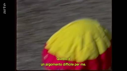 Un objet en tissu jaune et rouge est filmé en gros plan, accompagné d'un sous-titre en italien disant 'Questo è un argomento difficile per me'.