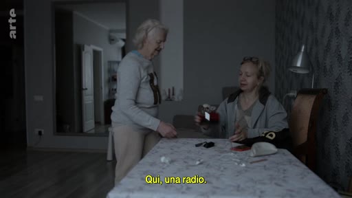 Deux femmes sont assises à une table dans un salon, discutant de la radio, comme l'indique le sous-titre à l'écran. L'ambiance est intimiste et domestique.