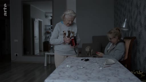 Deux femmes, dont une personne âgée, sont assises à une table dans un salon sombre et discutent en manipulant un petit appareil électronique, probablement une radio. L'ambiance est intimiste et domestique, illustrant une scène de vie quotidienne.