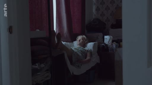 Une femme âgée est allongée sur un fauteuil dans une chambre faiblement éclairée, évoquant une atmosphère de nostalgie ou de solitude. Cette image illustre le thème de la mémoire et du temps qui passe dans un cadre domestique.