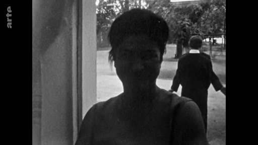 Une femme est filmée en gros plan dans un espace extérieur, tandis qu'une autre personne se tient de dos en arrière-plan. Cette séquence semble issue d'un document d'archive en noir et blanc.