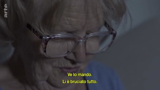 Une femme âgée, portant des lunettes, s'exprime face caméra lors d'un entretien filmé. Elle évoque des souvenirs ou des documents, comme l'indiquent les sous-titres en italien : « Ve lo mando. Lì è bruciato tutto. »
