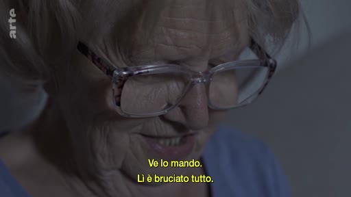Une femme âgée, portant des lunettes, s'exprime face caméra lors d'un entretien. Elle évoque des souvenirs ou des documents, comme l'indiquent les sous-titres en italien : 'Ve lo mando. Lì è bruciato tutto'.