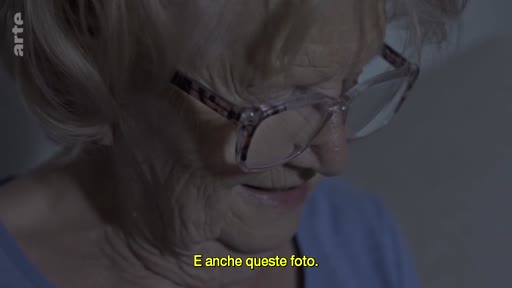 Une femme âgée portant des lunettes regarde des photographies tout en discutant, dans une séquence illustrant le souvenir et la transmission familiale.