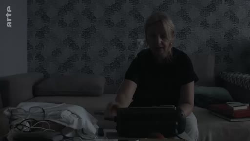 Une femme est assise sur un canapé dans un salon sombre et manipule un lecteur de cassettes audio pour écouter des enregistrements anciens.