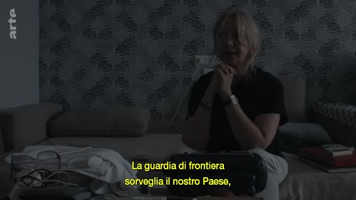Une femme blonde est assise dans une pièce sombre, écoutant une cassette audio, avec une sous-titre en italien évoquant la garde-frontière.