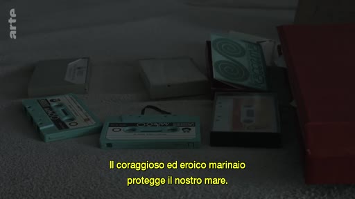 Plusieurs cassettes audio vintage sont disposées sur une surface, accompagnées de sous-titres en italien évoquant un marin héroïque.