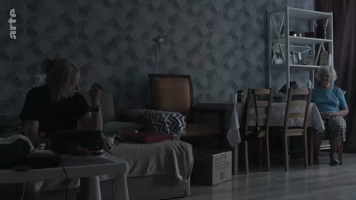 Une femme est assise près d'une table basse tandis qu'une personne âgée est assise à une table à manger dans un salon à l'éclairage tamisé, évoquant une ambiance de vie quotidienne ou de témoignage.