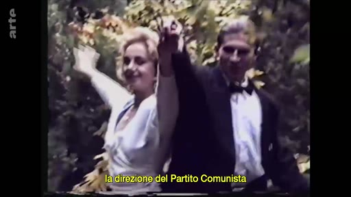 Un homme en costume et une femme en robe blanche saluent la foule, illustrant la direction du Parti Communiste. Cette image d'archive montre une scène de célébration politique.