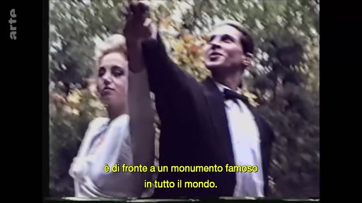 Un homme en smoking et une femme en robe blanche posent ensemble devant un monument célèbre, dans une séquence d'archives. L'image est accompagnée d'un sous-titre en italien évoquant un monument mondialement connu.