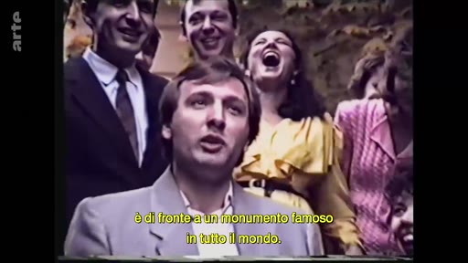 Un groupe de personnes, dont un homme au premier plan, s'exprime face caméra lors d'une interview en extérieur. Une légende en italien est visible à l'écran, évoquant un monument célèbre.