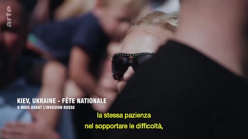 Une femme portant des lunettes de soleil est filmée lors de la fête nationale à Kiev, en Ukraine, six mois avant l'invasion russe. L'image est accompagnée d'un sous-titre en italien évoquant la patience face aux difficultés.