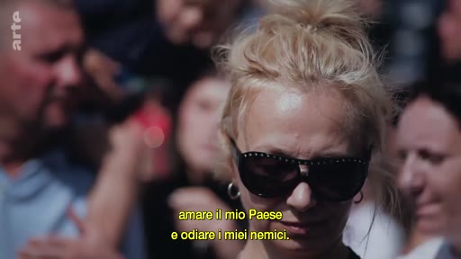 Une femme blonde portant des lunettes de soleil est filmée en gros plan au milieu d'une foule. Le sous-titre en italien affiche : 'amare il mio Paese e odiare i miei nemici'.