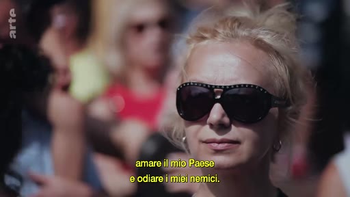 Une femme blonde portant des lunettes de soleil est filmée en gros plan lors d'une manifestation ou d'un rassemblement public. Le sous-titre à l'écran évoque des sentiments patriotiques et l'hostilité envers des ennemis.