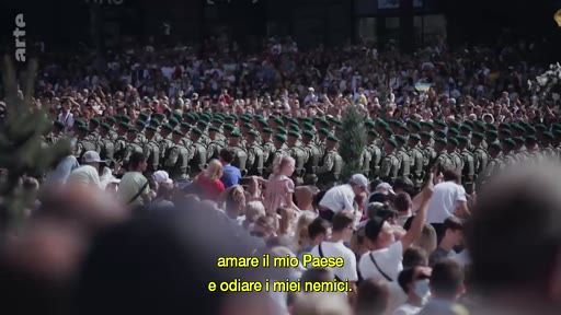 Une foule de spectateurs regarde un défilé de soldats en uniforme lors d'une manifestation publique, avec un sous-titre en italien évoquant l'amour du pays et la haine des ennemis.