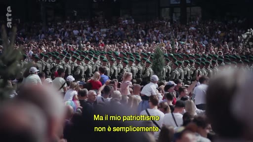 Une foule nombreuse regarde un défilé de soldats en uniforme lors d'une cérémonie publique, accompagnée d'un sous-titre sur le patriotisme.