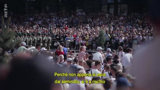 Une foule nombreuse assiste à un défilé militaire en extérieur, avec des soldats en uniforme marchant en rangs. Le sous-titre à l'écran souligne les risques liés à la dissidence ou au fait de sortir du cadre établi.