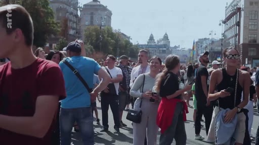 Une foule de civils marche dans une rue de Marioupol, en Ukraine, lors d'une journée ensoleillée. Les personnes sont filmées en plan d'ensemble dans un contexte de vie quotidienne urbaine.