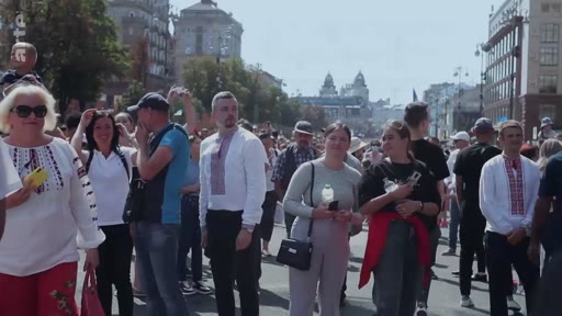 Un groupe de personnes, dont plusieurs portent des chemises traditionnelles ukrainiennes appelées vyshyvankas, participe à un rassemblement en extérieur. La scène montre une foule diversifiée dans un espace urbain, reflétant une manifestation culturelle ou patriotique en Ukraine.