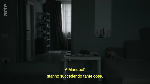 Vue intérieure d'une pièce sombre dans un appartement à Marioupol, accompagnée d'un sous-titre en italien évoquant la situation dans la ville. L'image illustre le quotidien dans une zone de conflit.