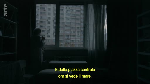 Une silhouette se tient devant une fenêtre dans un intérieur sombre, observant un grand immeuble d'habitation à Marioupol. Le sous-titre italien évoque la transformation du paysage urbain après les destructions de la guerre.