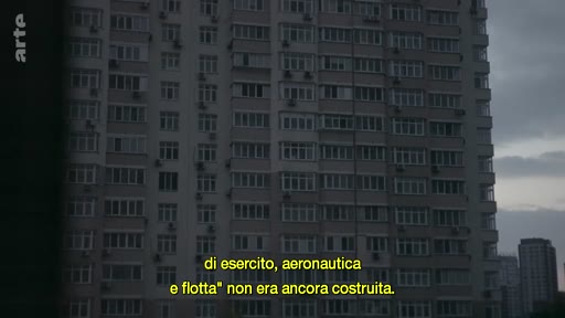 Vue extérieure d'un grand immeuble résidentiel à Marioupol, illustrant le contexte de la guerre en Ukraine. Les sous-titres en italien évoquent les structures militaires et la construction.