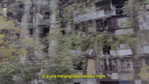 Vue extérieure d'un immeuble d'habitation en ruines à Marioupol, témoignant des destructions causées par la guerre en Ukraine. Le sous-titre ironique souligne le contraste entre la réalité dévastée et la vue sur la mer.
