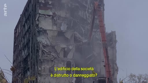 Une grue de chantier démolit un immeuble résidentiel fortement endommagé, illustrant les conséquences des destructions urbaines. L'image porte une question en italien sur l'état de l'édifice.