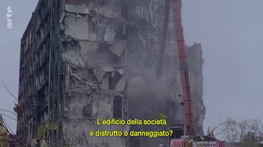 Une grue de chantier procède à la démolition d'un immeuble résidentiel fortement endommagé, probablement suite à un conflit armé. L'image illustre les conséquences de la guerre sur les infrastructures urbaines et le processus de nettoyage des décombres.