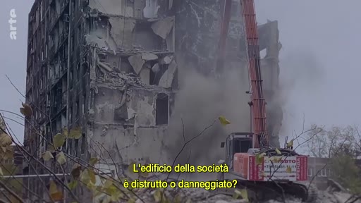 Une pelleteuse de chantier procède à la démolition d'un immeuble gravement endommagé, illustrant les conséquences de la guerre ou d'une catastrophe urbaine. L'image porte une question en italien sur l'état de destruction du bâtiment.