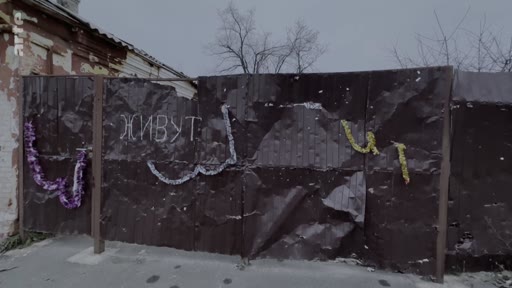 Une clôture en métal froissé et endommagée dans un environnement urbain, avec le mot russe « ЖИВУТ » (ils vivent) écrit en lettres blanches, symbolisant la résilience ou la survie dans une zone de conflit.