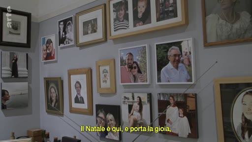 Un mur intérieur orné d'une collection de portraits de famille et de souvenirs photographiques encadrés, illustrant une atmosphère domestique et personnelle. La présence de sous-titres en italien suggère un contexte narratif lié à des souvenirs ou à une histoire familiale.