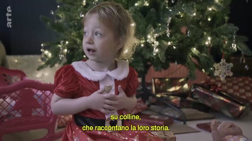 Une petite fille vêtue d'un costume de Père Noël s'exprime devant un sapin de Noël illuminé, dans une ambiance festive. Des sous-titres en italien apparaissent à l'écran.
