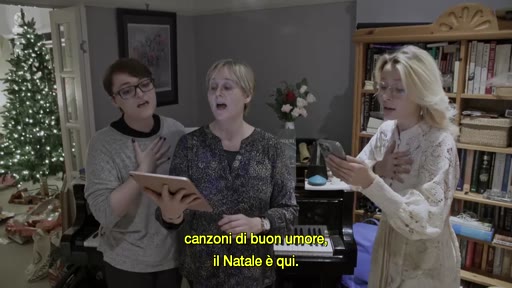 Trois femmes chantent des chants de Noël dans un salon décoré pour les fêtes, avec un sapin de Noël illuminé en arrière-plan. Elles tiennent des tablettes et des téléphones pour lire les paroles, illustrant une ambiance familiale et festive.