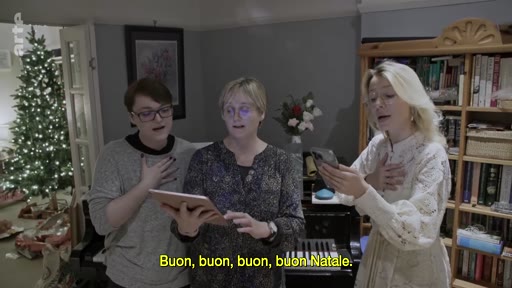Trois femmes chantent des chants de Noël dans un salon décoré pour les fêtes, avec un sapin de Noël en arrière-plan. Elles utilisent une tablette et un smartphone pour suivre les paroles, illustrant une scène de célébration familiale.