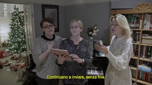 Trois femmes chantent ensemble dans un salon décoré pour Noël, avec un sapin illuminé en arrière-plan. Elles utilisent une tablette et un smartphone pour suivre les paroles, illustrant une scène de célébration familiale.