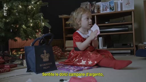 Une jeune enfant vêtue d'une robe rouge festive joue au pied d'un sapin de Noël, avec un sac portant l'inscription Buckingham Palace posé à ses côtés. La scène évoque l'ambiance des fêtes de fin d'année dans un contexte lié à la famille royale britannique.